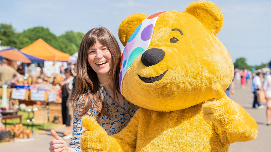 Natasha and Pudsey in a close-up shot smiling to camera with thumbs up. The antiques fair is visible in the background with people milling about in the sunshine