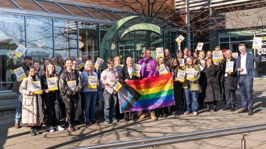 Dozens of Gateshead Pride organisers holding leaflets and the Pride flag as they smile for the camera.