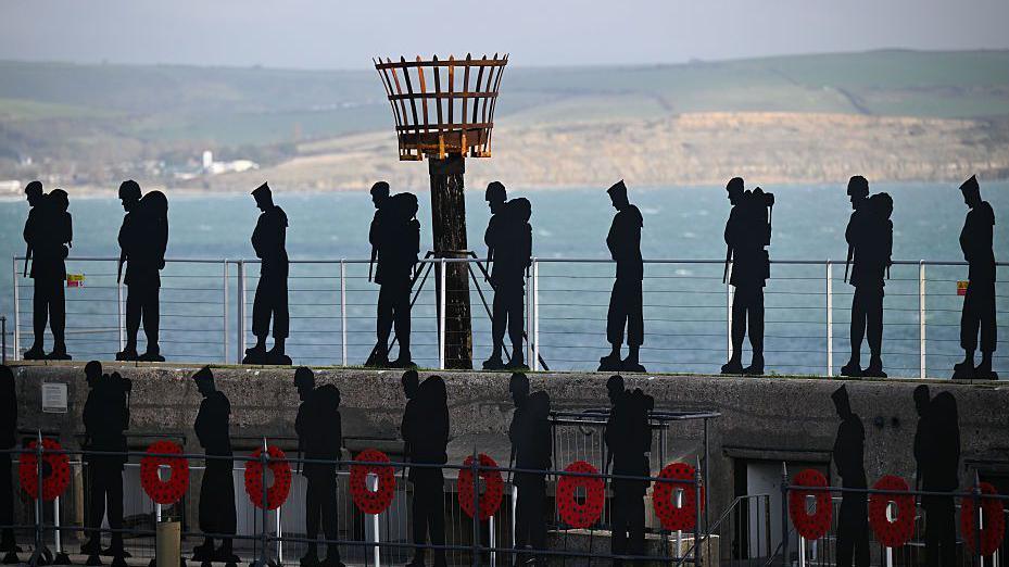 Life-size silhouetted figures of armed forces personnel line and stand atop a stone wall overlooking the ocean. The gate below is decorated with poppy wreaths.