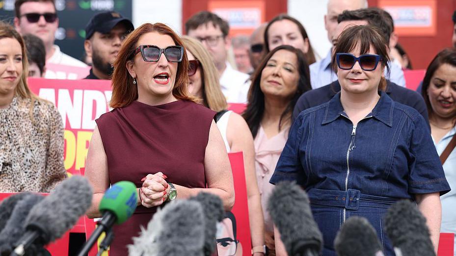 Louise Haigh, a woman with shoulder length ginger hair and sunglasses, addresses the crowd at Andy Burnham's by-election victory rally, as Anneliese Midgley, in a blue denim dress and sunglasses, looks on