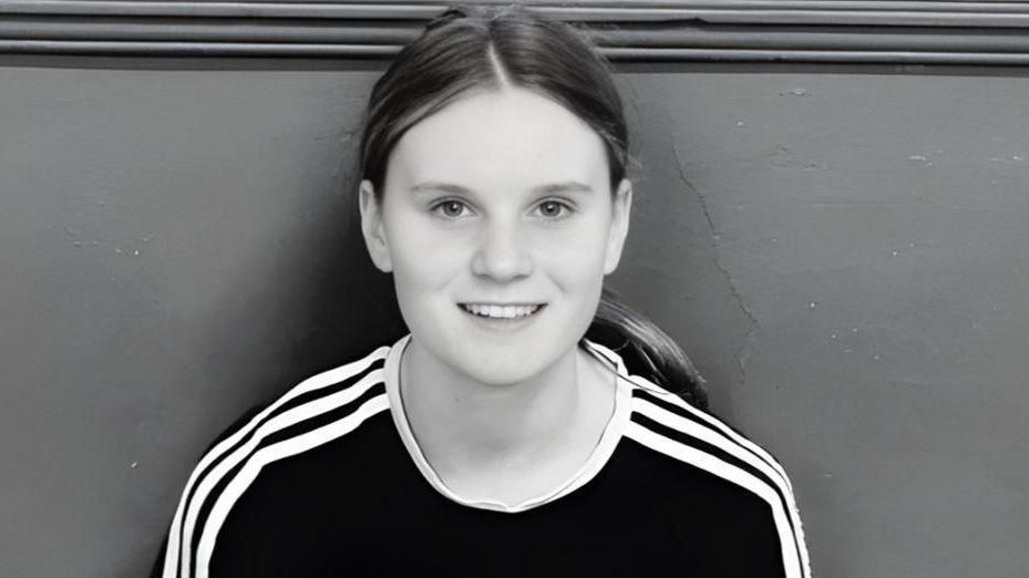 A black and white photograph of Holly Newton who is wearing a black top with white piping. She has her hair tied back and is smiling.