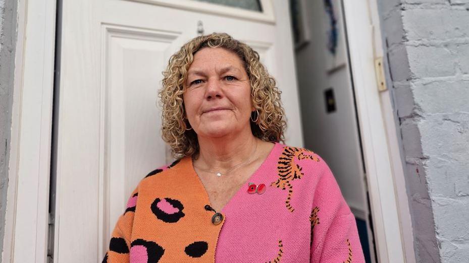 Angie Davies, who is wearing an orange and pink cardigan and has blonde curly hair, stands in front of an open door at the front of a property. 