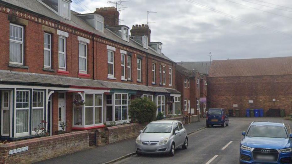 A row of red brick terraced housing on the left of the picture, with a silver and a purple car parked on the left hand side of the street with a metallic light blue car parked on the right
