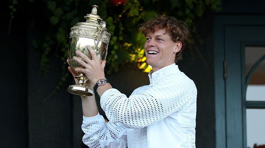 Jannik Sinner of Italy shows The Gentlemen's Singles Trophy to spectators from a balcony of Centre Court. He is smiling and holding the trophy up. He is wearing a white jacket and a watch with a gold face and black wrist strap. The trophy is gold.