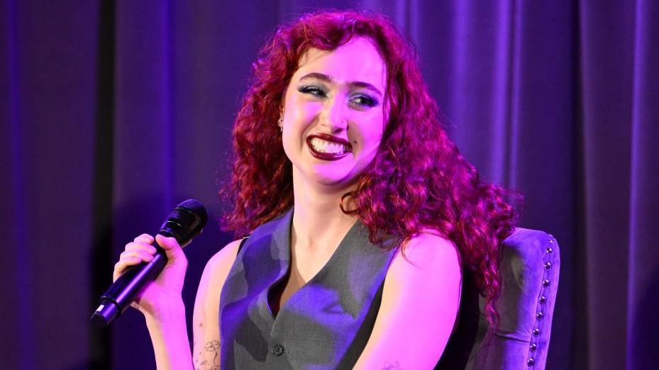 A woman with red hair sitting on a purple chair smiling, holding a microphone against a purple background