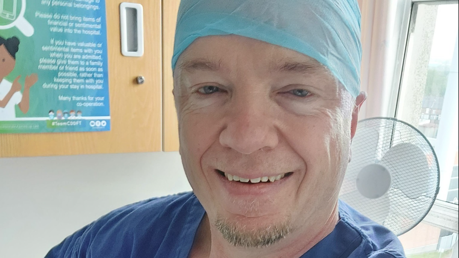 Joe Albon with blue eyes smiling at the camera. He is wearing blue hospital scrubs and a light blue protective covering over his head. 