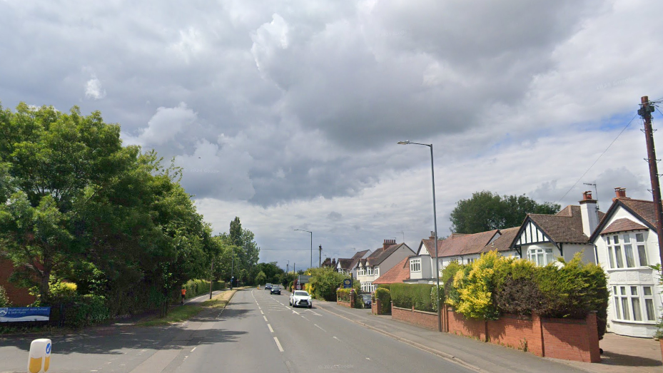 Alcester Road, a single lane highway with trees on either side and white houses on the right hand side.