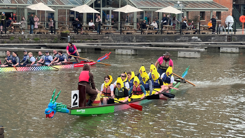 Two boats with 10 paddlers each are on the water in a city's docks, paddling towards the camera