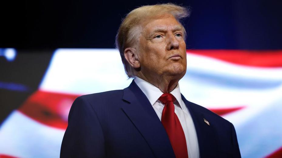 Donald Trump looking to the right of the camera. He wears a navy suit jacket, white shirt and red tie.