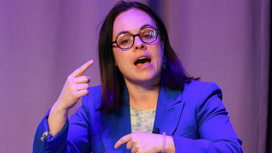 Kate Forbes, wearing small black round glasses and a blue blazer, she is mid speech and is gesturing with her index finger.