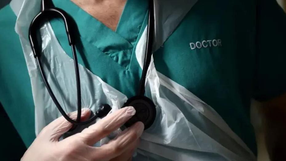 A close-up of the chest of a doctor who is wearing a green vest with the word doctor embroidered onto it. He is wearing white plastic gloves and a white plastic apron and has a black stethoscope hanging around his neck.