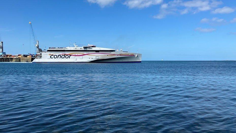 A white ferry with the Condor logo on the side in blue leaves a harbour on a sunny day.