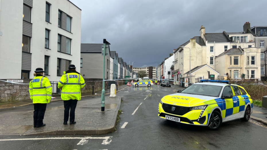 Two police officers in yellow standing on a pavement on the left. There is a parked police car on the right with a police cordon in the distance.