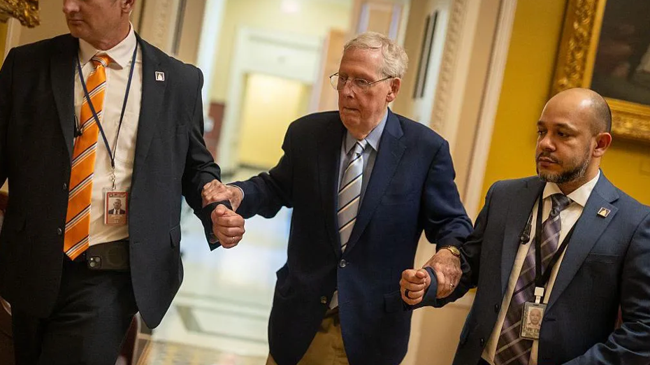 Mitch McConnell, wearing reading glasses and a blue suit jacket, leans on the arms of two men also wearing suits 