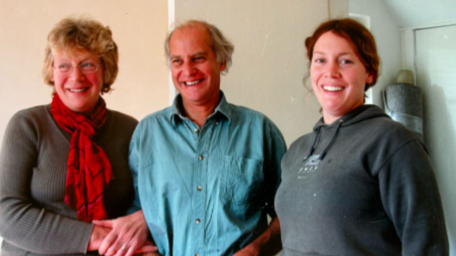 A family photo with Ros on the left, Martin in the middle and Debbie on the right. They are standing together with Ros and Martin arm in arm. 
Ros is in a grey jumper with a red scarf, Martin is wearing a blue shirt and Debbie is wearing a grey jumper. All are smiling at the camera. 