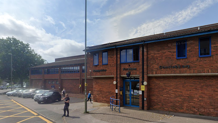 A Google Street view image of Fareham Police station. It appears to be summer as someone standing outside is wearing a t-shirt. The 1980s, two-storey, red-brick building has royal blue window frames and doors and a shallow pitched roof. Seven cars are parked diagonally in a line along the left of the image.