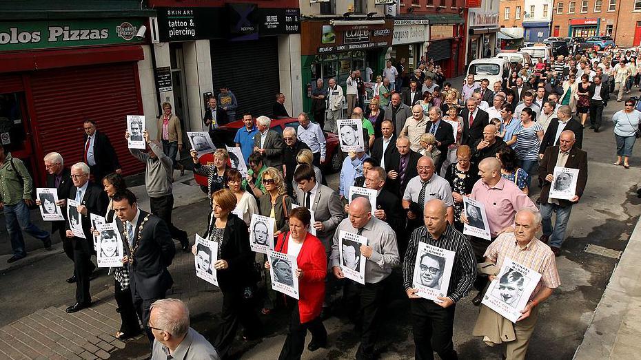 Families of the victims of Bloody Sunday carrying black and white images of their relatives, which is on white paper. They are dressed in a variety of coloured clothing as they walk past shops and restaurants on the streets of Londonderry.