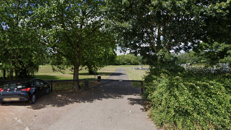 A green field beyond a row of trees. There is a concrete path running through the field, with a black gate in front of it. There is a parking area behind the treeline, with a black car parked up.