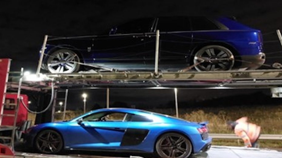 Two blue supercars are being towed on the back of a red vehicle on the side of a road. There is a grass verge and a barrier next to the vehicles.