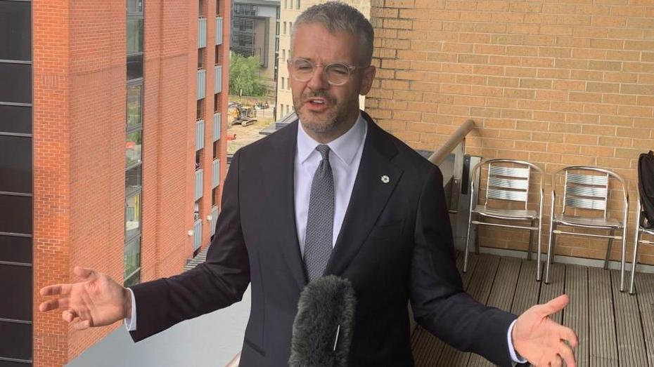 South Yorkshire Mayor Oliver Coppard speaks with his arms outstretched in front of a microphone.