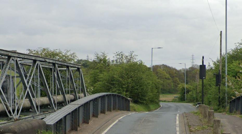 A road runs over a metal bridge. There are stone bollards on the right-hand side of the road. There is a traffic light and two streetlights in the distance. The road beyond the bridge has hedges on either side before the carriageway curves out of view.