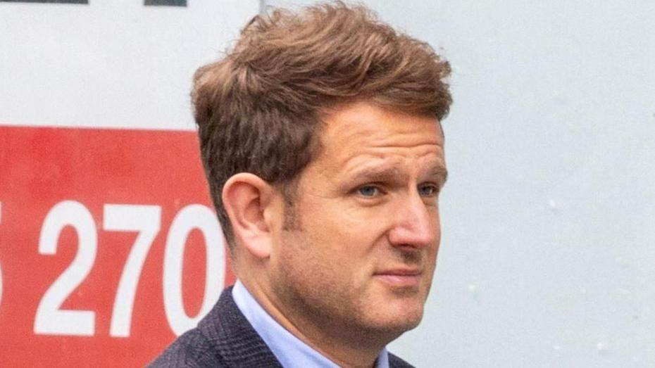 A man with brown hair, wearing a dark suit, looking away from camera