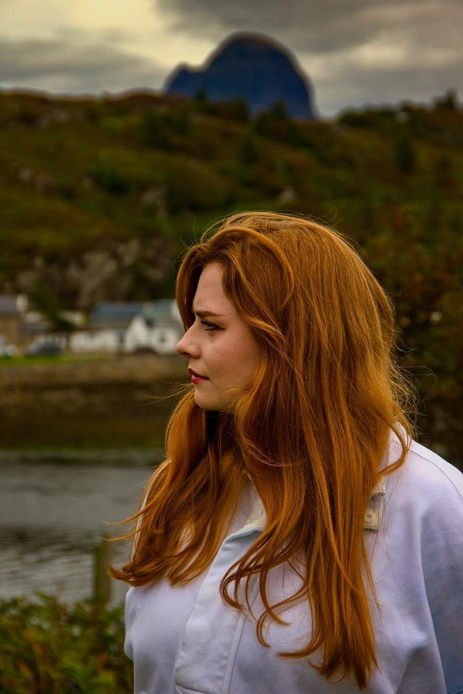 Leah McBain is a young woman with long ginger hair. She is standing outside in the village of Lochinver and is looking into distance with houses and a bay in the background.