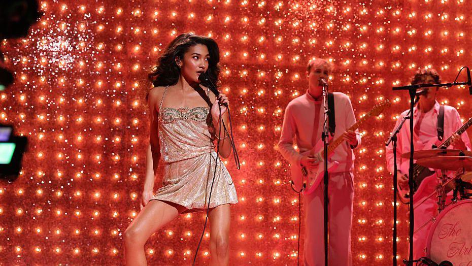 Olivia Dean performing on a red stage in a silver sparkly dress.