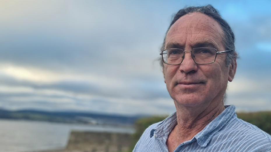 Professor Donald Nicholson stands on the coast with the sea and cliffs behind holding a copy of his book 'How to Set Up and Run a Law Clinic'. He's wearing a blue and white striped shirt, and metal glasses and looks at the camera from the right of the image. He's holding the book out to his right side. The book is teal and white with capitalised lettering. The sky in the background has a mix of cloud, blue sky and the sun shining through highlighting white clouds. 