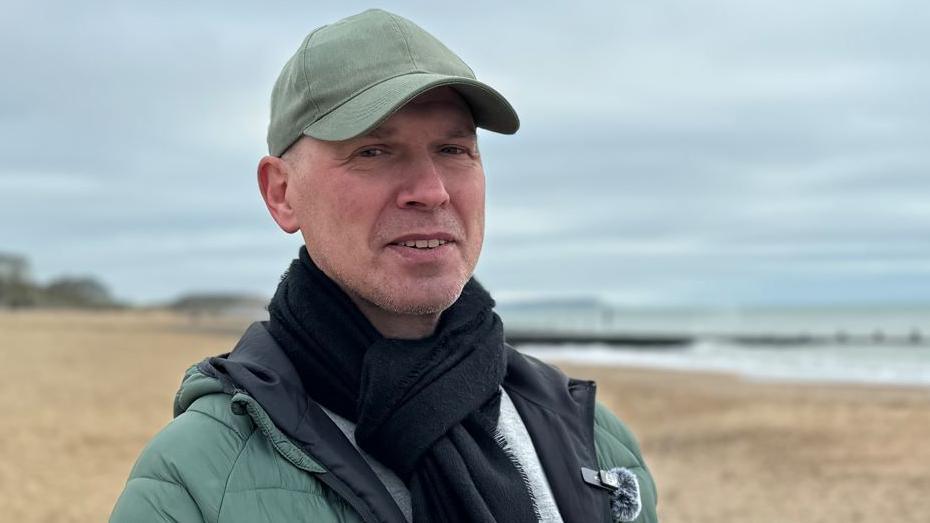 Jason is standing on a beach and wears a khaki green puffa jacket, black scarf and light khaki green baseball cap. He has light stubble on his face and smiles into the camera