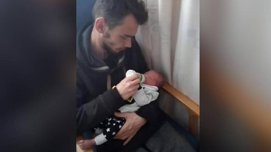 A man with brown hair holding a baby in his left arm. With his right hand, he is feeding the child with a bottle of milk. They are sitting in a chair with a curtain visible on the left of the pair of them.