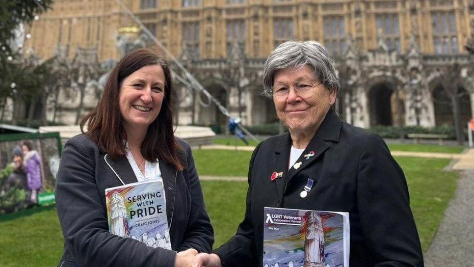 Two women, both wearing black blazers and holding books, shaking hands within the Palace of Westminster, on a lawn, with the bottom of Big Ben behind them