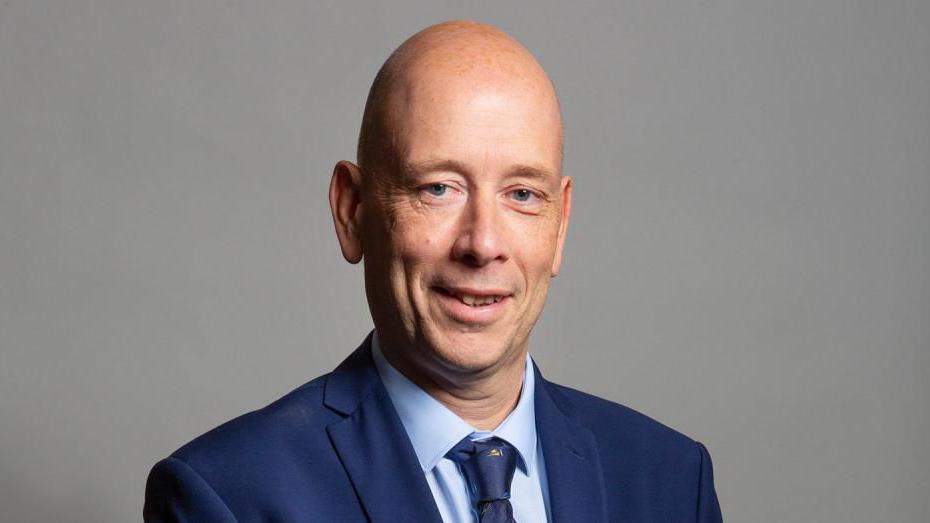 A bald man smiles at the camera. He is wearing a navy blue suit with a light blue shirt and navy and yellow tie.