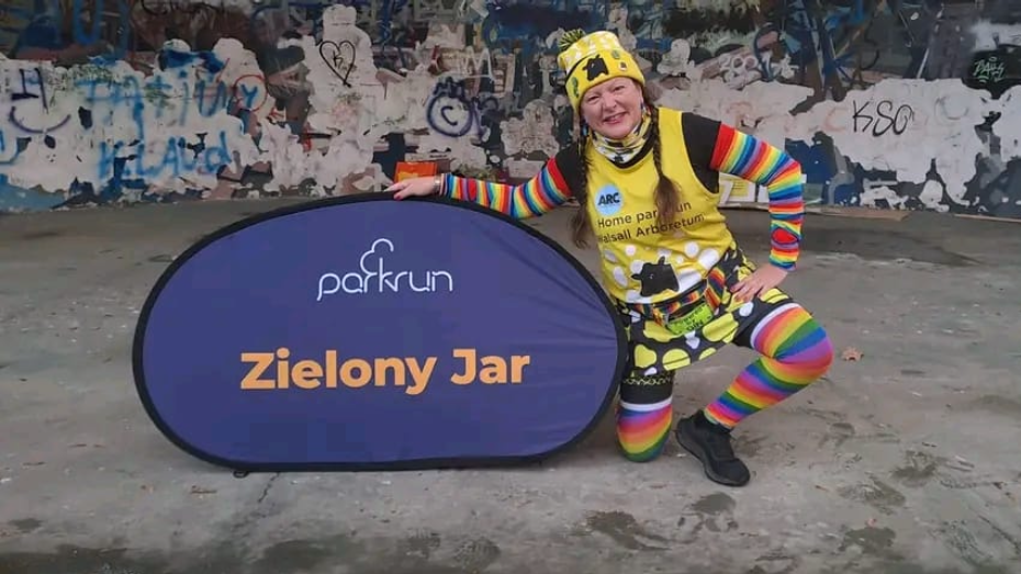 A woman wearing a yellow Parkrun vest and a multi-coloured striped t-shirt and leggings kneels in front of a purple Parkrun sign saying Zielony Jar, on a concrete floor in front of a graffitied wall