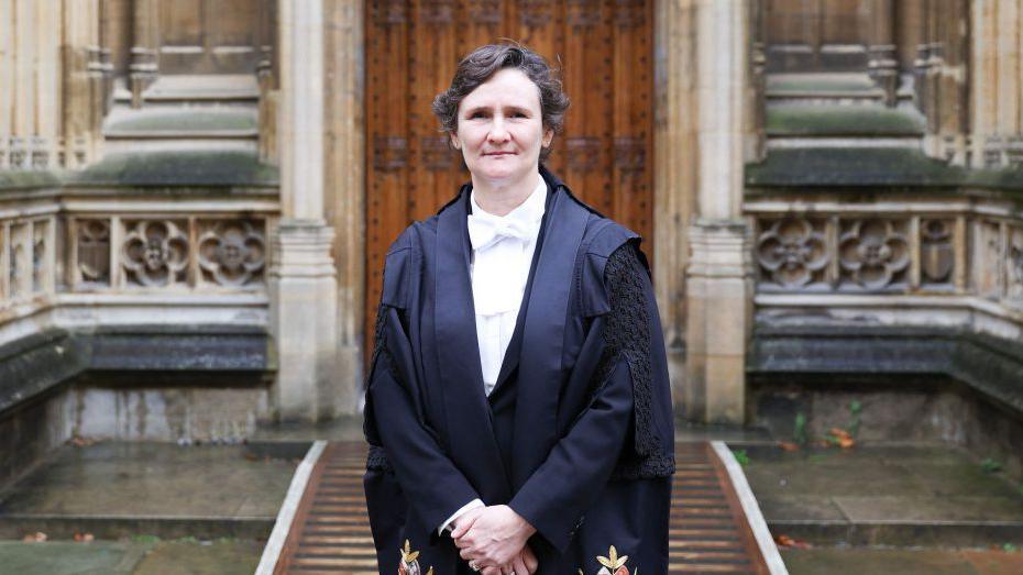 Prof Tracey in black gowns and a white shirt. She is stood with her arms down in front and has short brown hari. Behind her is a rather old looking stone doorway.