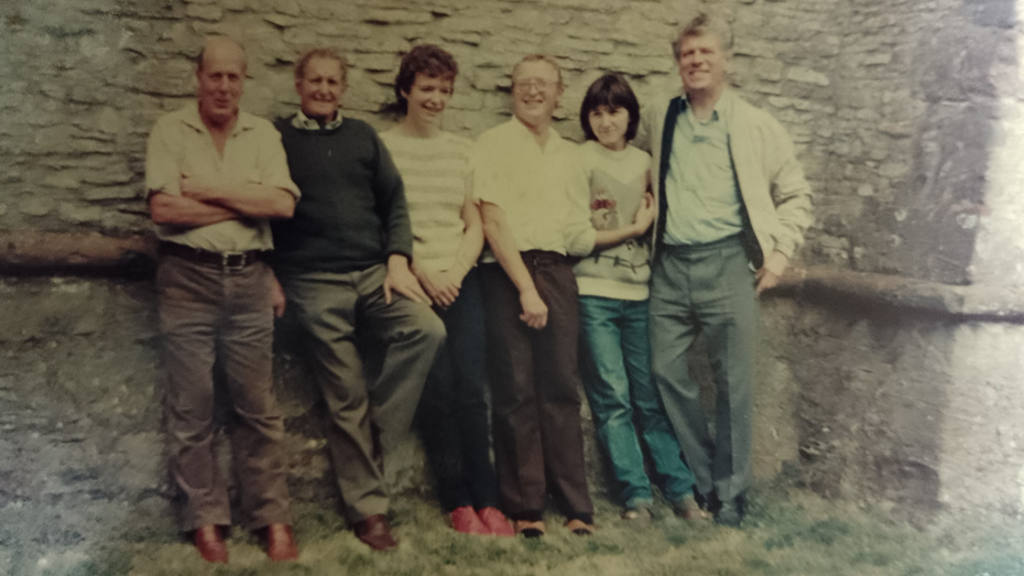 A very old, faded, photo of six bell ringers standing outside by a church tower in casual clothes and smiling to camera