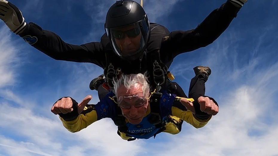 Brain, who has grey hair and wears and yellow and blue boiler suit, gives two thumbs up to the camera as he skydives. He is attached in tandem to an instructor, who is wearing all black.