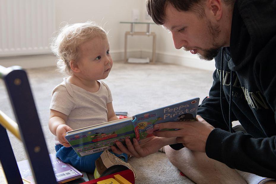 Callum Fay, sitting on a cream carpet on the floor, reading a Peppa Pig book to his son Archie, who is staring at his father, totally rapt.