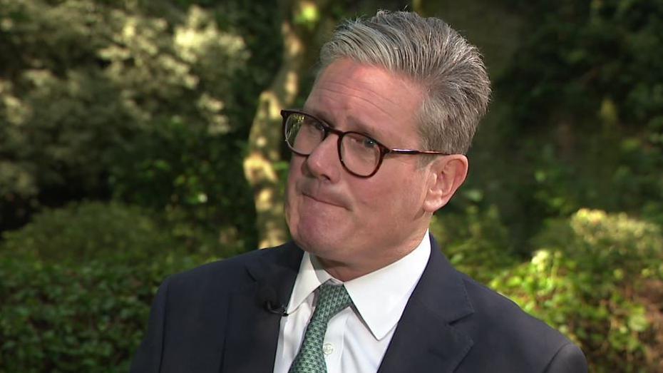 Sir Keir Starmer, wearing a dark suit, a white shirt and a patterned green tie, talks to the BBC's Tim Iredale, with greenery in the background