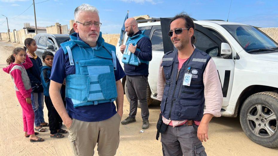 David Anderson and another man are standing in a sandy environment. David wears a blue bullet proof vest and the other man wears a gilet which identifies him as an aid worker. Three children stand in the background.