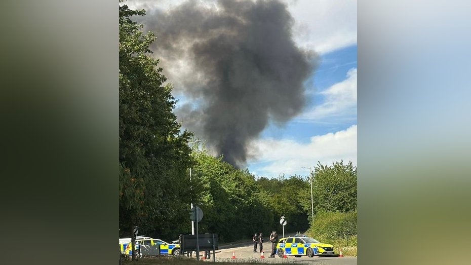 Large plume of black smoke rises from behind trees, in the foreground are a number of police cars