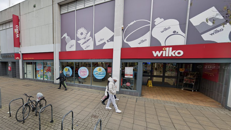 A 2 storey store with a red sign which says 'wilko' on the front. The bottom floor is glass fronted. In front is a wide pavement and bike racks. Some people are walking past.