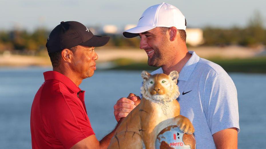 Host Tiger Woods shakes hands with winner Scottie Scheffler at the 2024 Hero World Challenge event, beside the tournament trophy