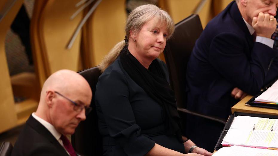 Shona Robison, wearing her hair in a ponytail and a black scarf and jackets sitting in the Scottish Parliament chamber. To her left is John Swinney.