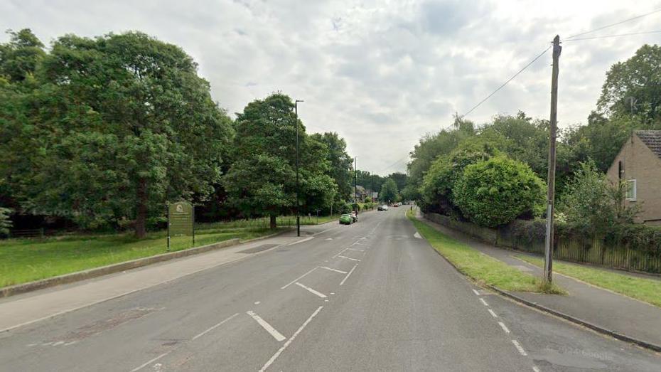 A Google Streetview image of Wetherby Road in Leeds. The road stretches off into the distance. On one side of the there are trees and grass and on the other side a telegraph pole and a brick-built bungalow.