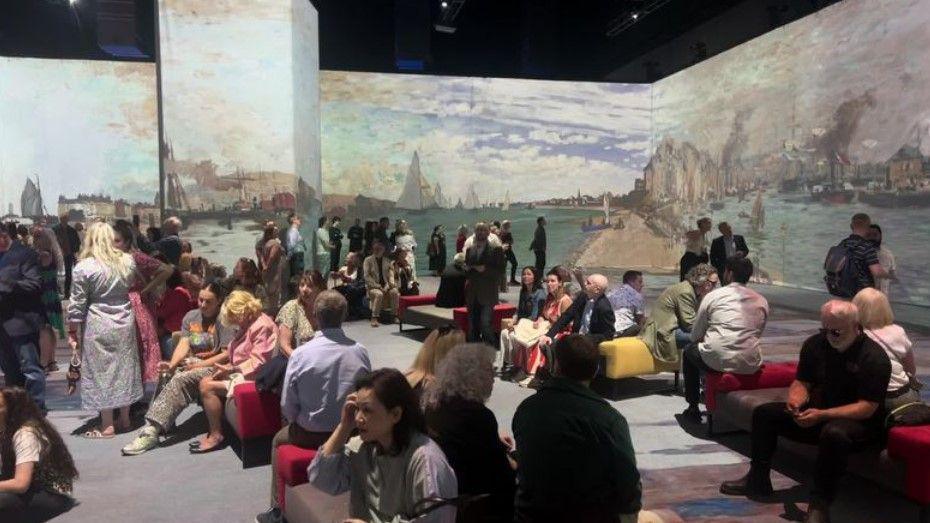 Many people sitting down to view projected images of Monet's Impressionist paintings, depicting ships in a harbour.