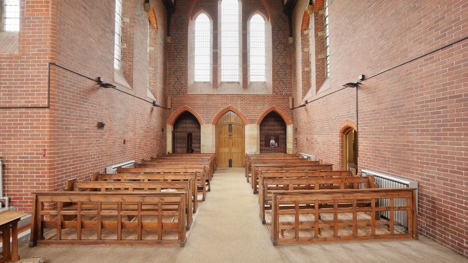 Rows of wooden benches sit either side of a chapel with an aisle down the middle. At the end is a large wooden door underneath three large-pained glass window frames