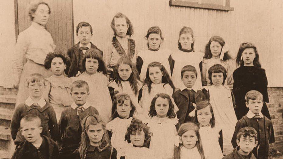 A black and white photo showing 23 young pupils outside a school and an adult woman stood next to them