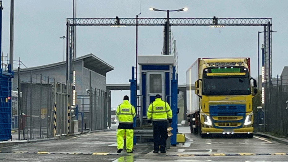 Officials await the arrival of an HGV at a border check area. They are both in yellow hi-vis. A lorry is coming through the port.