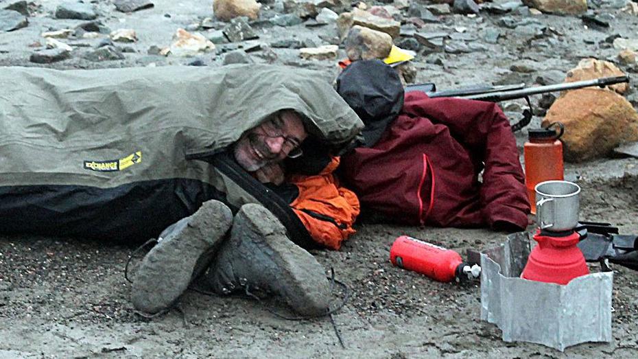 Prof John Marshall wild camping on rocky ground on an expedition to Spitsbergen in 2023. He smiles from a gap in his sleeping bag, which mostly covers his head. A pair of mud-encrusted boots, a small gas stove and other equipment are beside him.